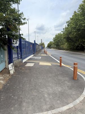 Newly reinstated footway with timber bollards, flush kerbs, and fresh double yellow lines curving past the fenced site frontage in Barking