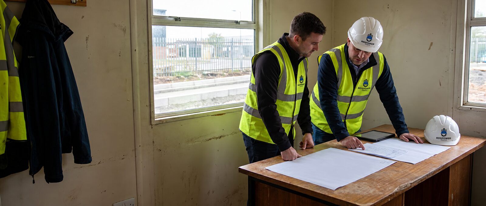 Two construction staff reviewing drawings and a tablet in a site cabin